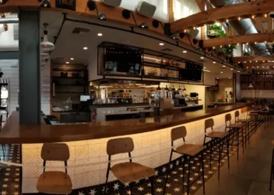 Panoramic view of a modern, well-lit restaurant interior featuring a long bar counter with barstools, wooden ceiling beams, and tables with chairs. A few patrons are seated at tables.