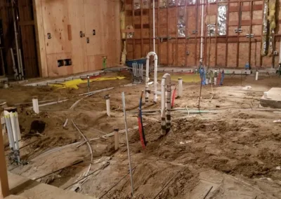 A room under construction with exposed wooden framing and plumbing pipes emerging from the ground. Electrical wiring and insulation are visible on the walls.