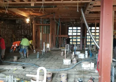 Construction workers performing various tasks inside a building framework with exposed wood and wiring, concrete floor under preparation with pipes and rebar visible.